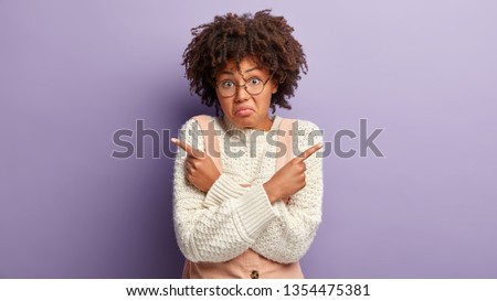 Photo of clueless dark skinned woman points fore fingers at upper corners, being hesitant, chooses between two objects, purses lips, has curly hair, poses over purple background. What to choose?