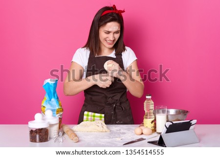 Happy housewife wears kitchen apron dirty with flour, white t shirt, red headband, kneads dough while having videocall with her husband. Studio picture of smiling baker isolated over pink background.