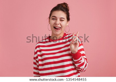 Portrait of a happy young woman with freckles, wears striped longslive, showing peace gesture and sticking her tongue out isolated over pink background.