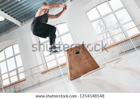 Side view of strong fitness man doing jump exercises on box in gym. Fit athlete wearing sportswear and jumping over box in sport hall