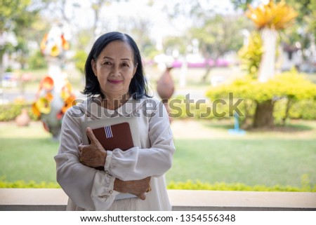 elderly retirement thai women standing by the window and laptop with notebook in hands by the garden background