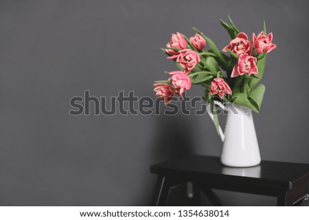 A bouquet of pink peony tulips in a white jug stand on a black bench.