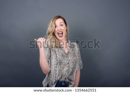 Omg concept. Stupefied caucasian female with surprised expression, opens eyes and mouth widely, points aside with thumb, shows something strange on gray background. Advertisement concept.
