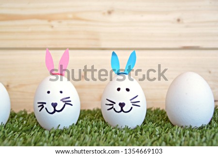 A row of white eggs lie on a short-cut lawn, one of the eggs has colorful bunny ears glued on and a rabbit's face painted - concept for Easter with wooden background with space for text