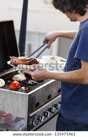 Early 30's caucasian man loading a plate with the cooked items from the Sausages, onion slices and pita bread that are getting ready on an outdoor barbecue grill.