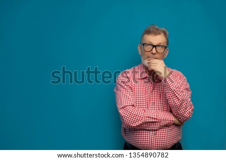 Thoughtful old man wearing smart casual shirt and stylish glasses dreaming about something posing on blue background.