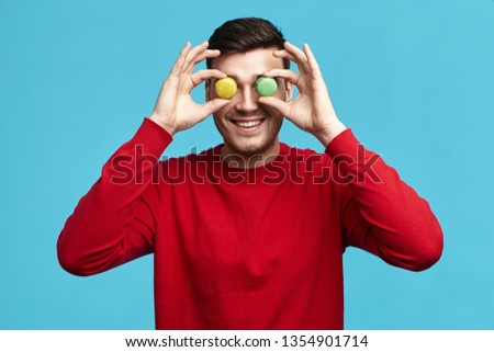 Food, pastry, joy and fun concept. Picture of funny unshaven young brunette male in red sweatshirt posing isolated at blue studio wall, holding two round almond cookies at his eyes and smiling happily