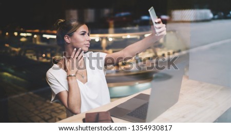 Attractive hipster girl dressed in casual look clicking selfie photos via cellular phone while sitting at table with modern laptop computer, charming woman taking pictures via application on cellphone