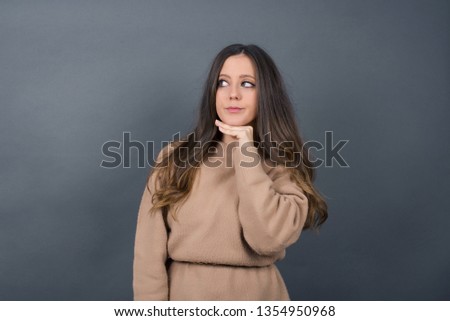 Dreamy  female with pleasant expression, wearing casual clothes, looks sideways, keeps hand under chin, thinks about something pleasant, poses against gray background.
