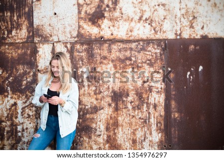 Millennial blonde beautiful girl use cellular phone to message or search on social media - urban abandoned wall in background - steel colors and portrait of nice people