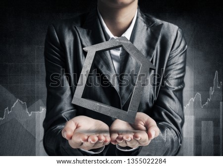 Closeup of businessman in black suit keeping stone house symbol in hands with sketches on dark wall on background. 3D rendering.