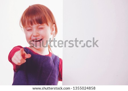 Funny little child girl laughing behind a white board and pointing with her finger to you. Space for text.