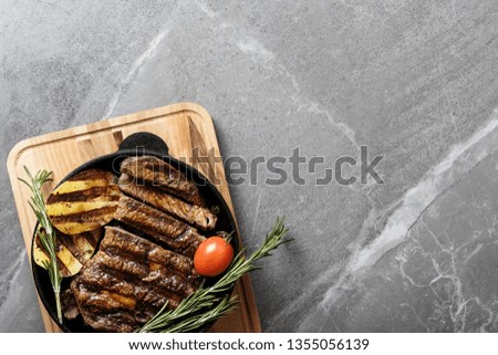 fried rib-eye steak in pan with cherry tomatoes, fried potatoes and green sprigs of rosemary on gray marble table top view, grilled meat on barbecue flat lay