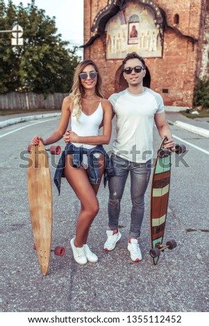 Beautiful young couple man and woman walk summer city, hands skateboards, longboard. Happy smiling posing having fun. Walk on weekend love relationships strong family. Emotions positive and caring.