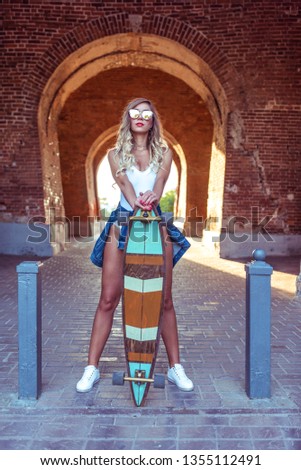 Beautiful woman stands in brick arch. Girl in white body with jeans, summer in the city. Concept style, fashion, fashion. Skateboard longboard, sunglasses, brick wall on background.