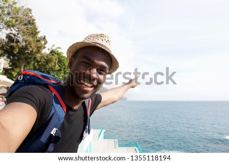 Portrait of smiling african american travel man taking selfie by the sea and pointing finger
