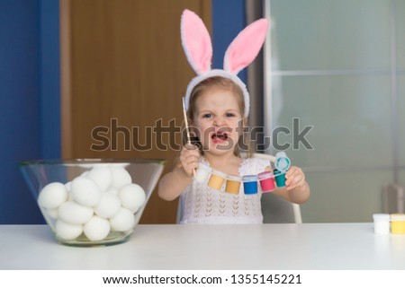 Beautiful little blonde girl, has happy fun smiling face, pretty eyes, white t-shirt, hare ears, paint easter eggs. Child portrait and kids hobby concept. Holiday accessories. 