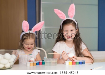 Beautiful little girls sisters, has happy fun smiling face, pretty eyes, white t-shirt, hare ears, paint easter eggs. Child portrait and kids hobby concept. Holiday accessories. 