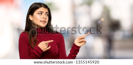 Teenager girl with turtleneck frightened and pointing to the side at outdoors
