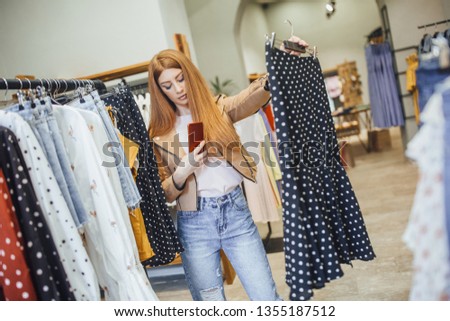 A young redhead girl in a women's clothing store photographs a skirt on her mobile phone. online shopping