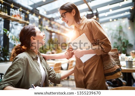 Excited surprised young lady in glasses holding laptop and touching shoulder of friend while greeting her in cafe, girl bumping into friend in public place Royalty-Free Stock Photo #1355239670