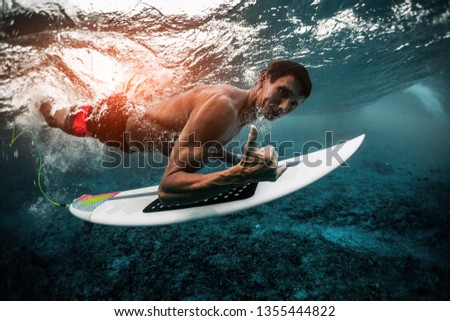 Muscular adult surfer dives under the wave with surfboard and shows the Shaka sign
