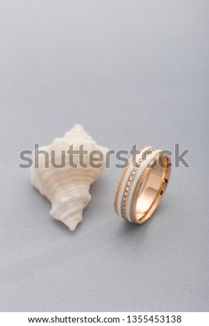 Gold ring with white enamel and diamonds on gray background with seashell. Minimalist concept. Original wedding ring band with gemstones