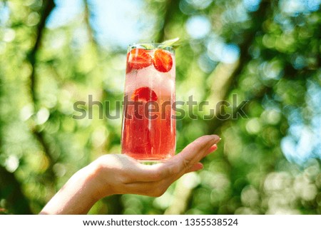 Woman holding in hand fresh strawberry lemonade with ice and mint in glass with green background outdoors, copy space. Cold summer drink. Sparkling glass with berry cocktail