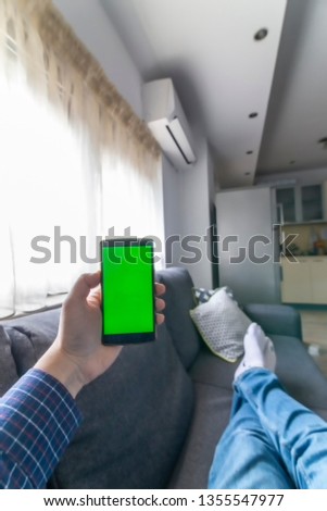 Person laying on a couch with smartphone in his hand and controlling the air conditioner. Blank green chroma screen.