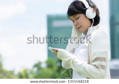 Happy asian woman closing her eyes and listening to music on mobile phone. Blur building background.