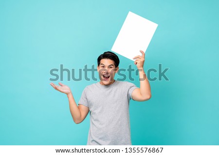 Portrait of a young cheerful asian male in  excited gesture promoting a blank placard with his arms raised in blue studio background