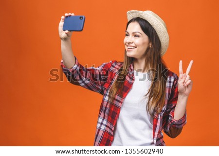 Young cheerful attractive brunette is smiling on the orange background. She is taking selfie on the camera of her phone, wearing casual summer outfit and a hat. 