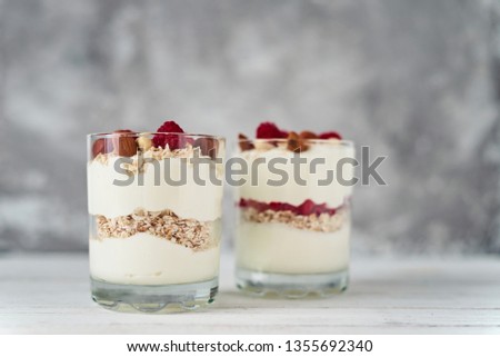 Two glasses of greek yogurt granola with raspberries, oatmeal flakes and nuts on white background. Healthy nutrition