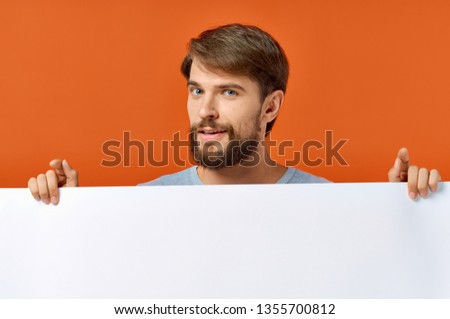 Cute bearded man holding in front of him a large white drawing paper on an orange background