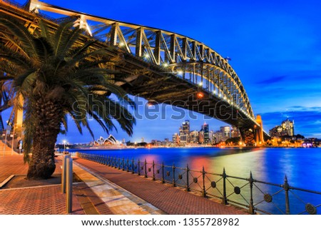 Side-view of Steel arch of the Sydney Harbour bridge during blue hour at sunset . The Opera house is tiny, far away from being the main point and can't be removed from overall cityscape. Royalty-Free Stock Photo #1355728982