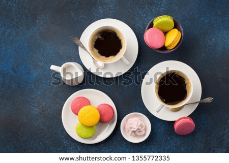 White cups of black coffee, served on saucer with macaroons biscuits, meringue and cream. Branch over blue wooden background. Flat lay.