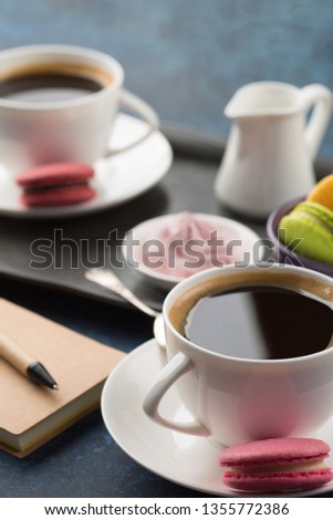 White cups of black coffee, served on saucer with macaroons biscuits, meringue and cream. Branch on blue wooden background, selective focus