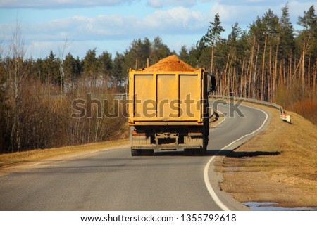 Transportation of bulk building materials, heavy overloaded dump truck carries sand on a European countryside one way asphalt serpentine road turn on pine forest and blue sky background at autumn day Royalty-Free Stock Photo #1355792618