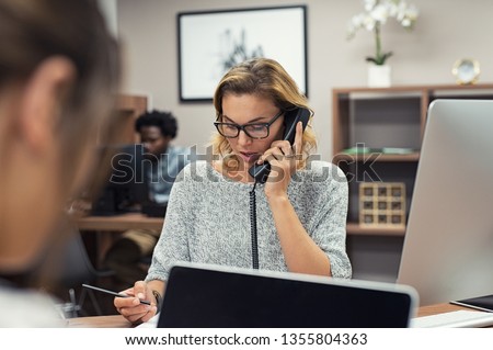 Mature businesswoman talking on phone in office. Casual business woman talking on landline phone in a creative agency. Beautiful blonde receptionist with eyeglasses taking client details over phone. Royalty-Free Stock Photo #1355804363