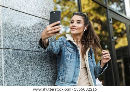 Beautiful young brunette woman wearing jacket, carrying backpack walking outdoors, taking a selfie
