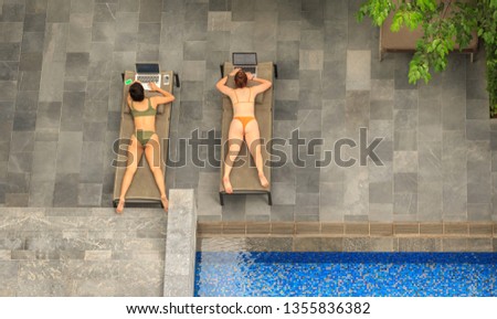 Top view of two girls in bikini typing on laptops while  lying by swimming pool