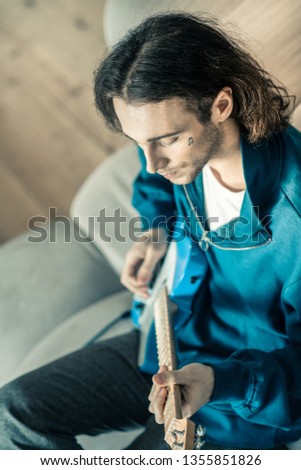 Bright electronic guitar. Thoughtful good-looking guitarist playing his musical instrument while resting on grey couch