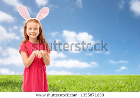 easter, holidays and childhood concept - happy red haired girl wearing bunny ears headband over blue sky and grass background