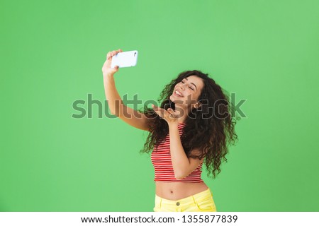 Photo of happy woman 20s wearing summer clothes smiling and using cell phone for selfie isolated over green background