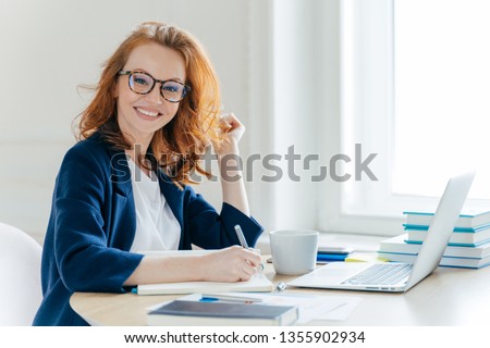 Sideways shot of prosperous businesswoman with foxy hair, smiles positively, records information in notepad, drinks coffee, searches information on laptop computer, develops strartup project Royalty-Free Stock Photo #1355902934