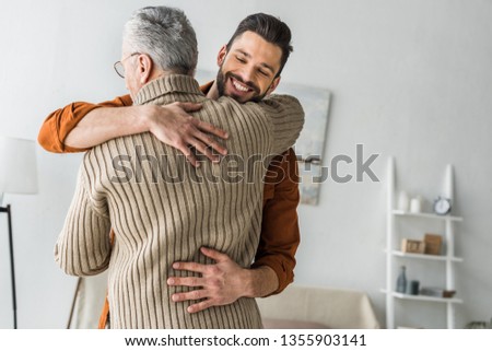 happy bearded man smiling while hugging elder father at home  Royalty-Free Stock Photo #1355903141