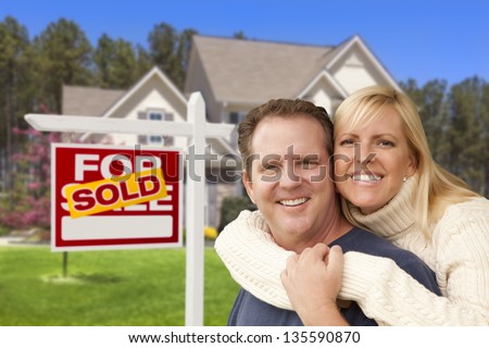 Happy Couple Hugging in Front of Sold Real Estate Sign and House.