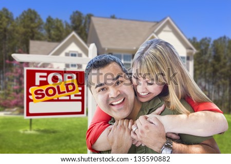 Happy Hugging Mixed Race Couple in Front of Sold Home For Sale Real Estate Sign and House.