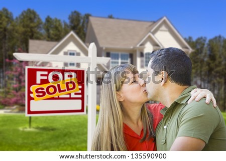 Happy Mixed Race Couple in Front of Sold Home For Sale Real Estate Sign and House.