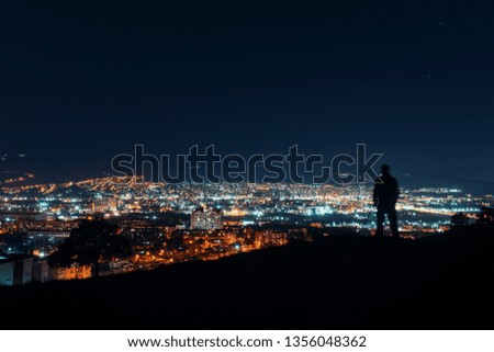 Night over big city. Photographer silhouette standing on top of the hill over the city, making night photography.